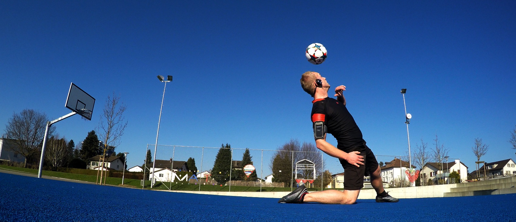 Aeroskiller kniet auf blauem Sportplatz und balanciert einen Fussball auf der Stirn — Panorama-Actionaufnahme unter klarem Himmel.